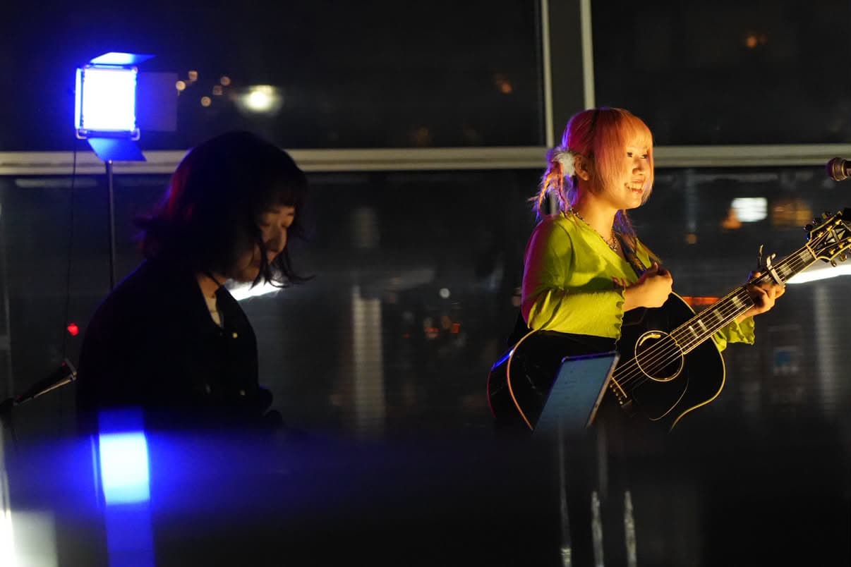 2026年10月20日 新浦安駅前でギターを弾きながら笑顔を見せる東田明莉(SHE IS LIGHT)と、サポートキーボードの大原杏慈友